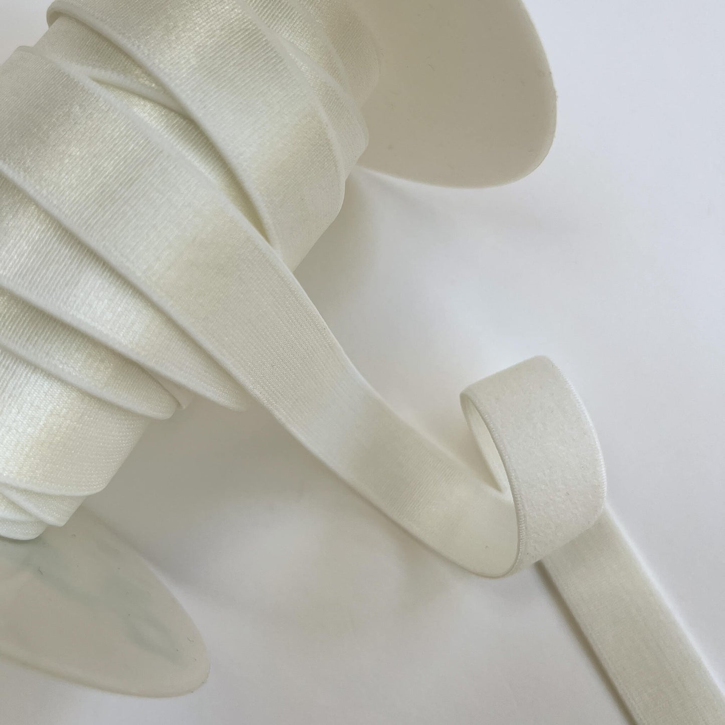 A close-up of a spool of wide, flat elastic ribbon in a warm off-white or ivory color, with a length of the ribbon unspooled on a plain light background.