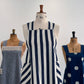Three vintage dress forms displaying different apron designs: a central cream apron with navy blue vertical stripes and striped pockets, flanked by a chambray apron with white geometric lace detail and a navy blue apron with white polka dots.