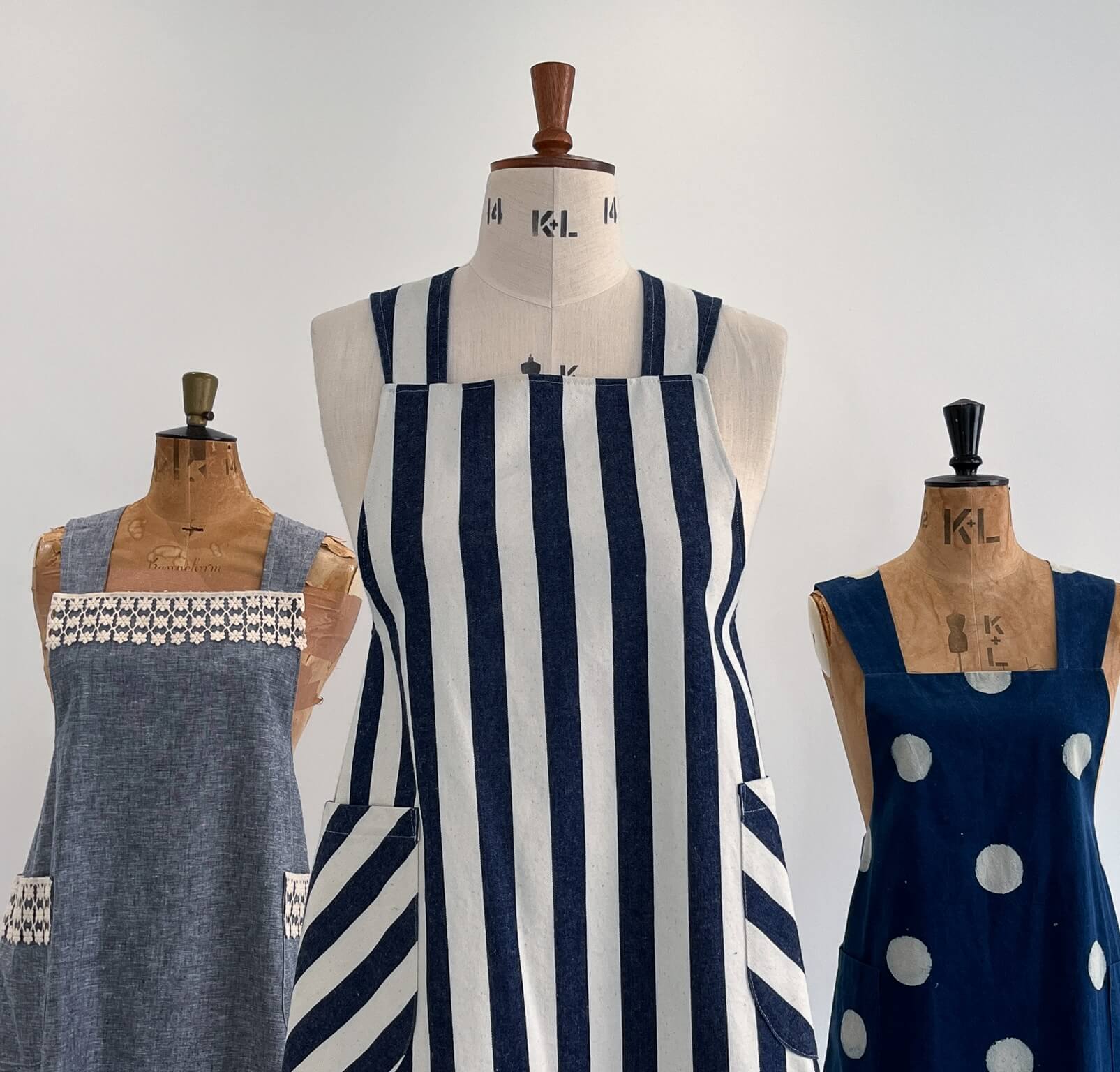 Three vintage dress forms displaying different apron designs: a central cream apron with navy blue vertical stripes and striped pockets, flanked by a chambray apron with white geometric lace detail and a navy blue apron with white polka dots.