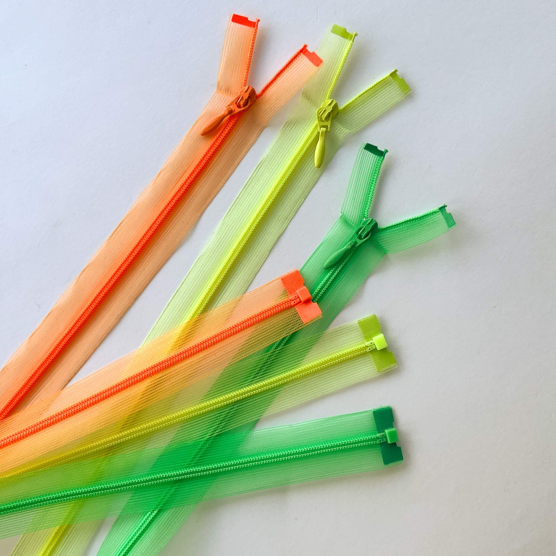 Couture Featherweight open ended zip - 100cm length, light weight mesh zippers in orange, yellow-green, and green, arranged diagonally on a light off-white background.