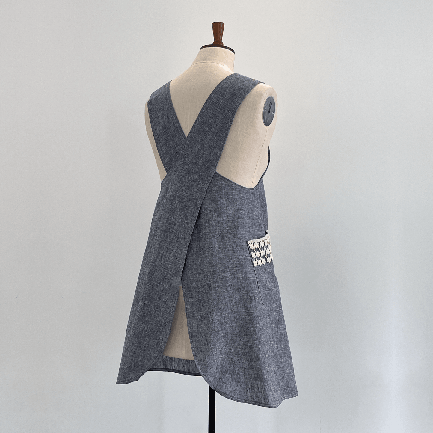 A chambray linen apron with a crossover back and a decorative lace detailed large pocket.