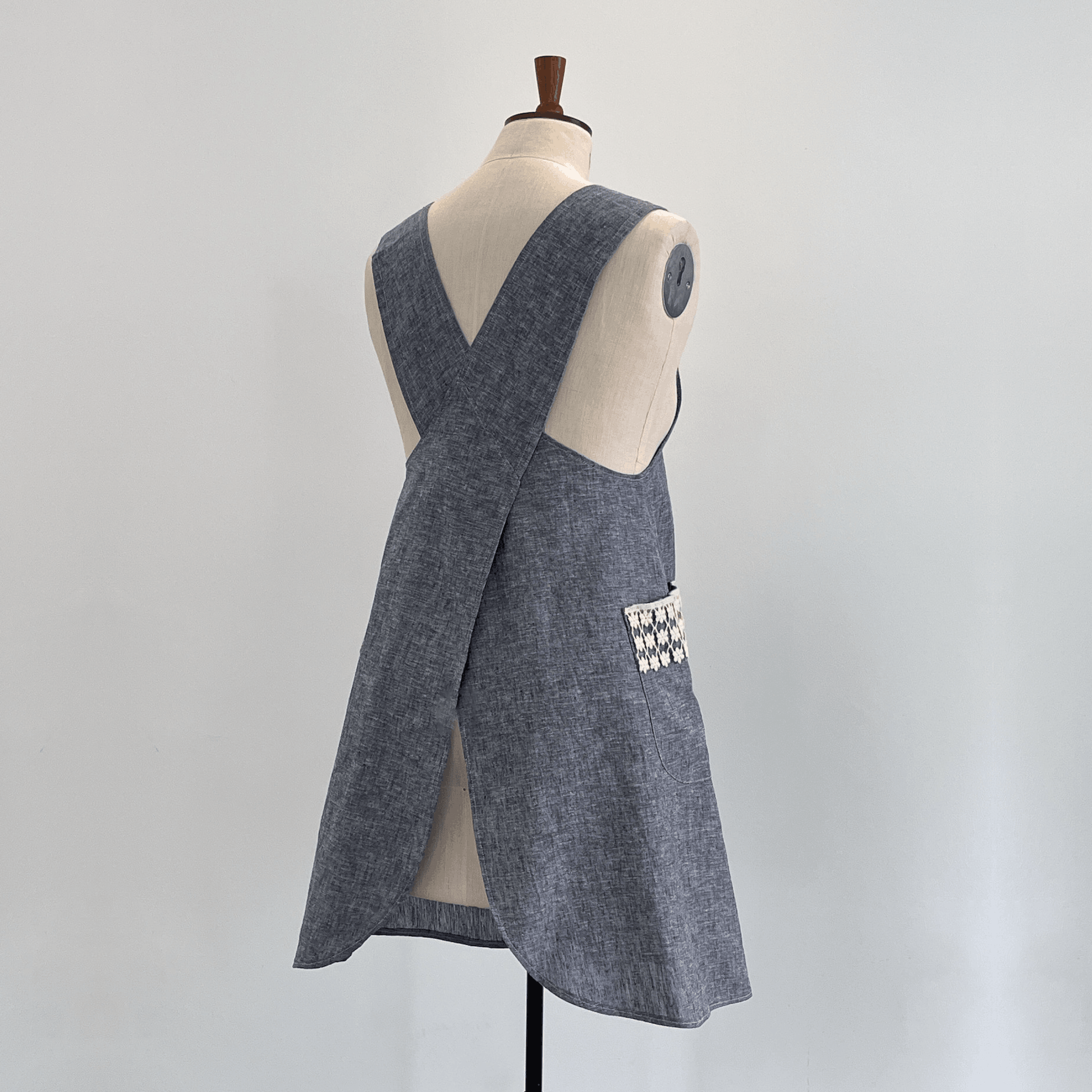A chambray linen apron with a crossover back and a decorative lace detailed large pocket.