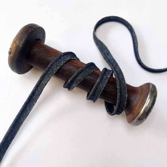 A close-up of a denim blue piping cord wrapped around a wooden spool with a silver metal cap, set against a plain white background.