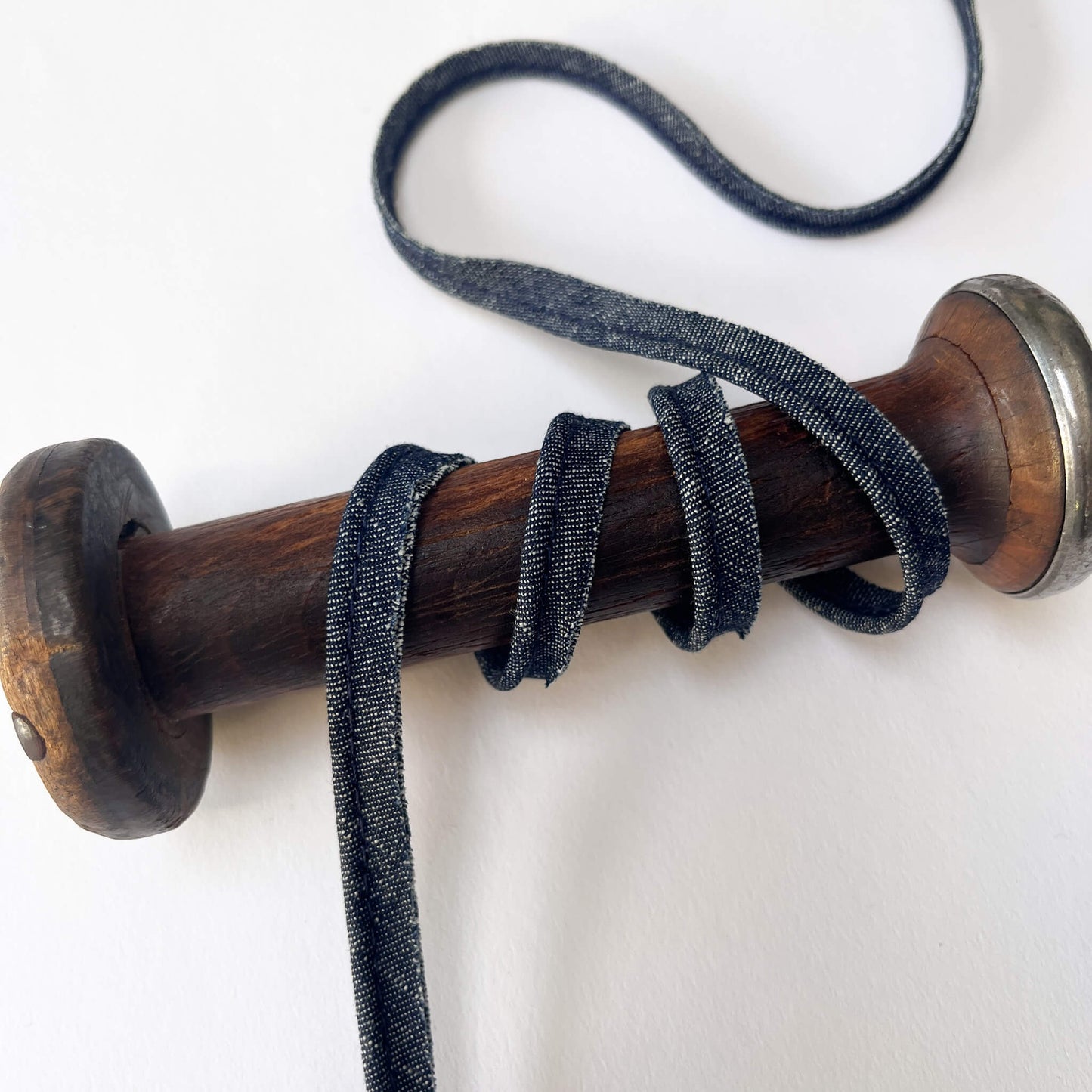 A close-up of a denim blue piping cord wrapped around a wooden spool with a silver metal cap, set against a plain white background.