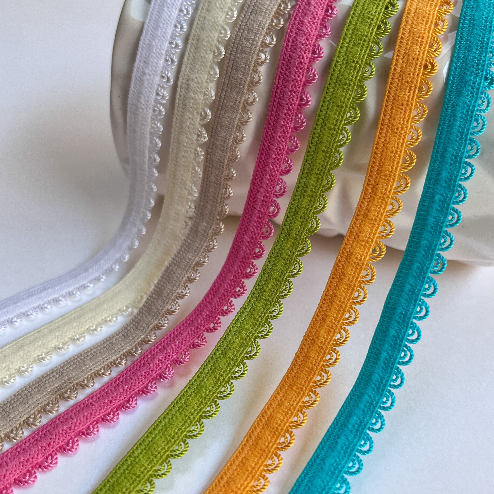 Six decorative elasticated picot lace trim ribbons in white, beige, pink, green, yellow, and turquoise, arranged diagonally on a white background.
