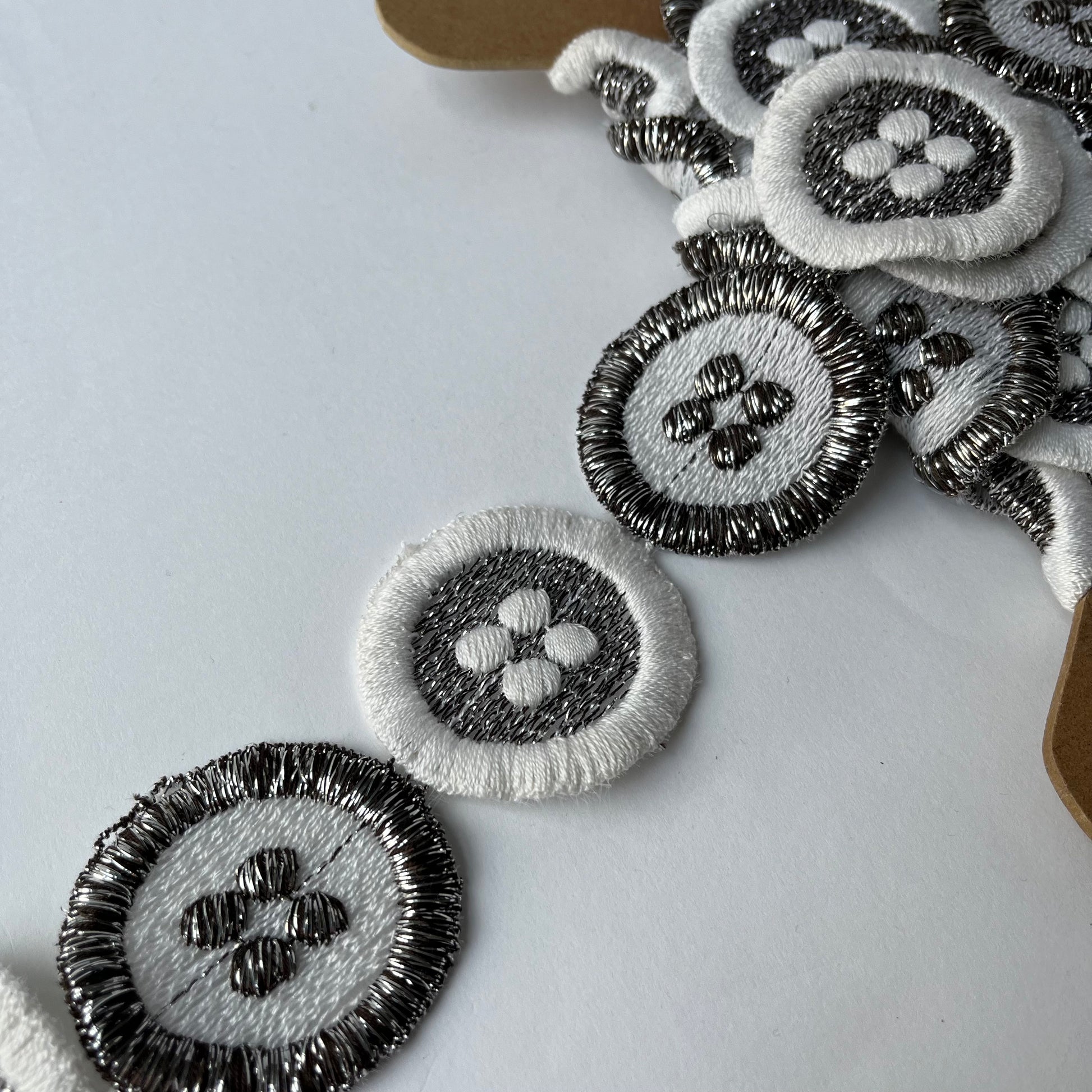 Close-up of embroidered oval patches with a floral or clover-like center motif, shown in black, white, and gray thread on a light background.