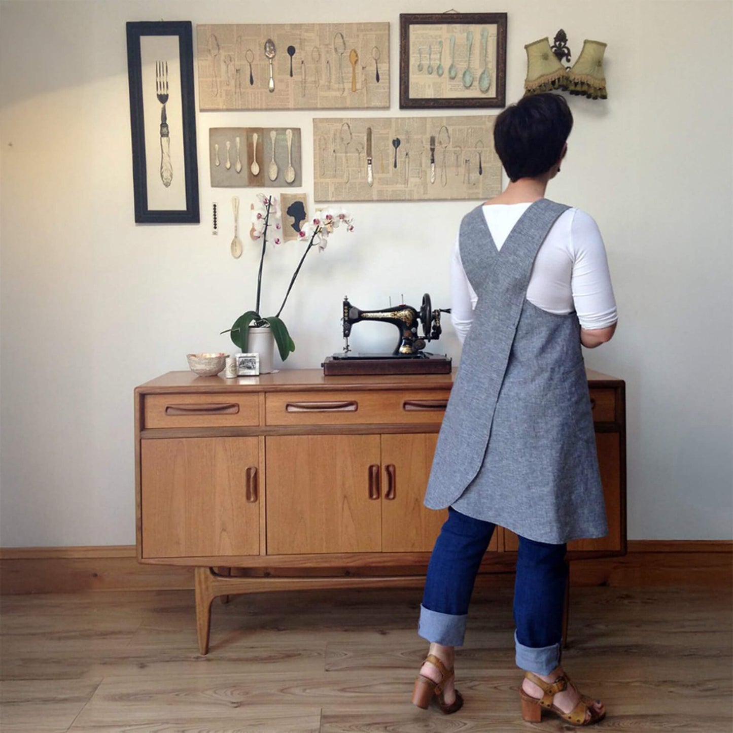 A woman viewed from behind wearing a linen apron over a white t-shirt and blue jeans, standing in a vintage room in front of a wooden sideboard.