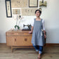 A woman wearing a gray linen apron over a white long-sleeve top and blue jeans stands in a room with a wooden mid-century modern cabinet, vintage sewing machine, and framed utensil art on the wall.