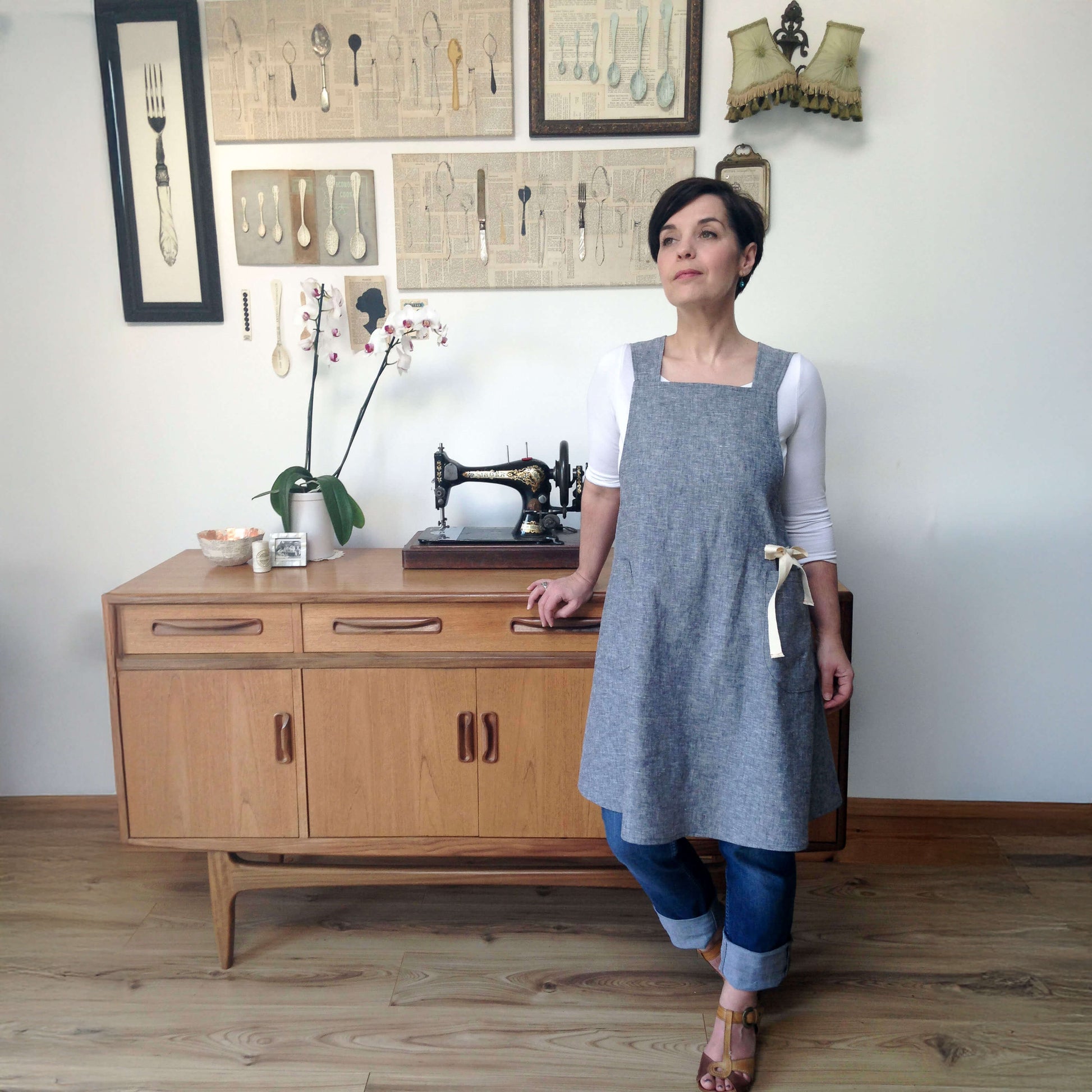 A woman wearing a gray linen apron over a white long-sleeve top and blue jeans stands in a room with a wooden mid-century modern cabinet, vintage sewing machine, and framed utensil art on the wall.