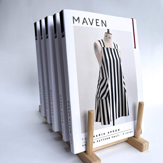 A stack of The Maria Apron sewing patterns displayed on a light wood stand against a clean white background. 