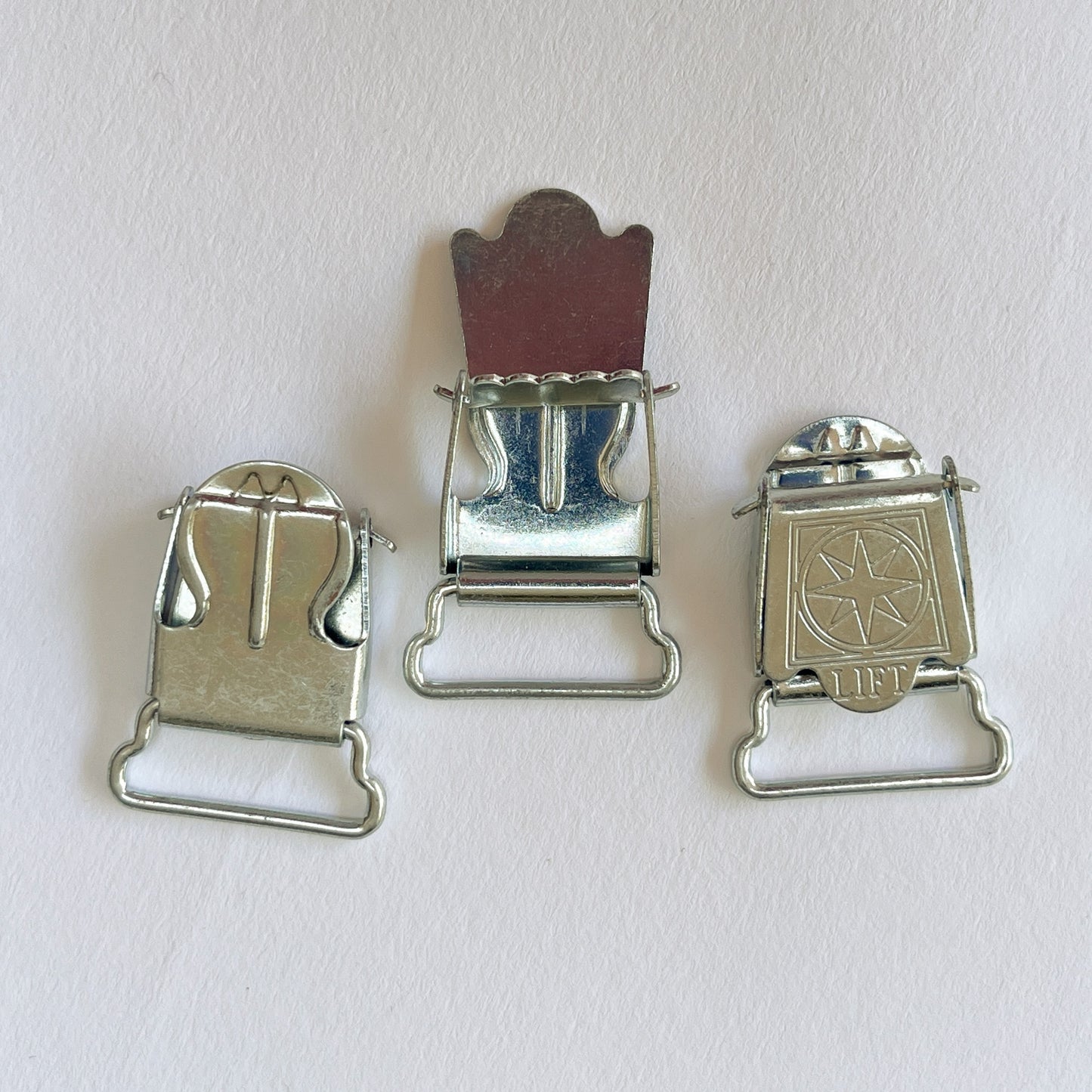 Small silver metal suspender clips or clasps with a star-and-circle emblem on the front, photographed on a plain light background. Can also be used for Braces.