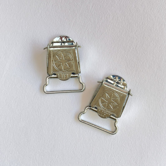 Two small silver metal suspender clips or clasps with a star-and-circle emblem on the front, photographed on a plain light background. Can also be used for Braces.